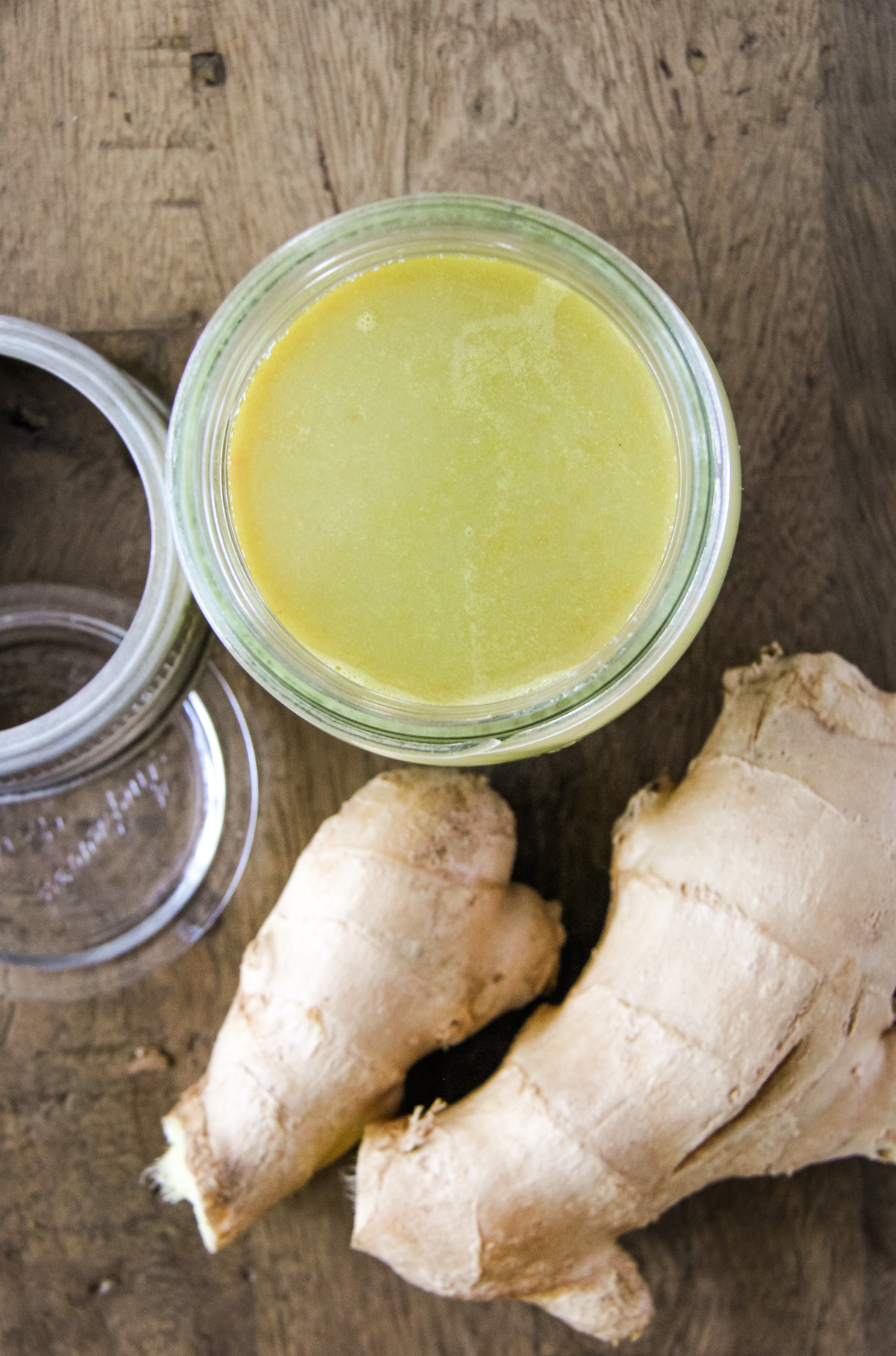 a top down image of a jar of fresh ginger juice. a fresh root sits in the foreground
