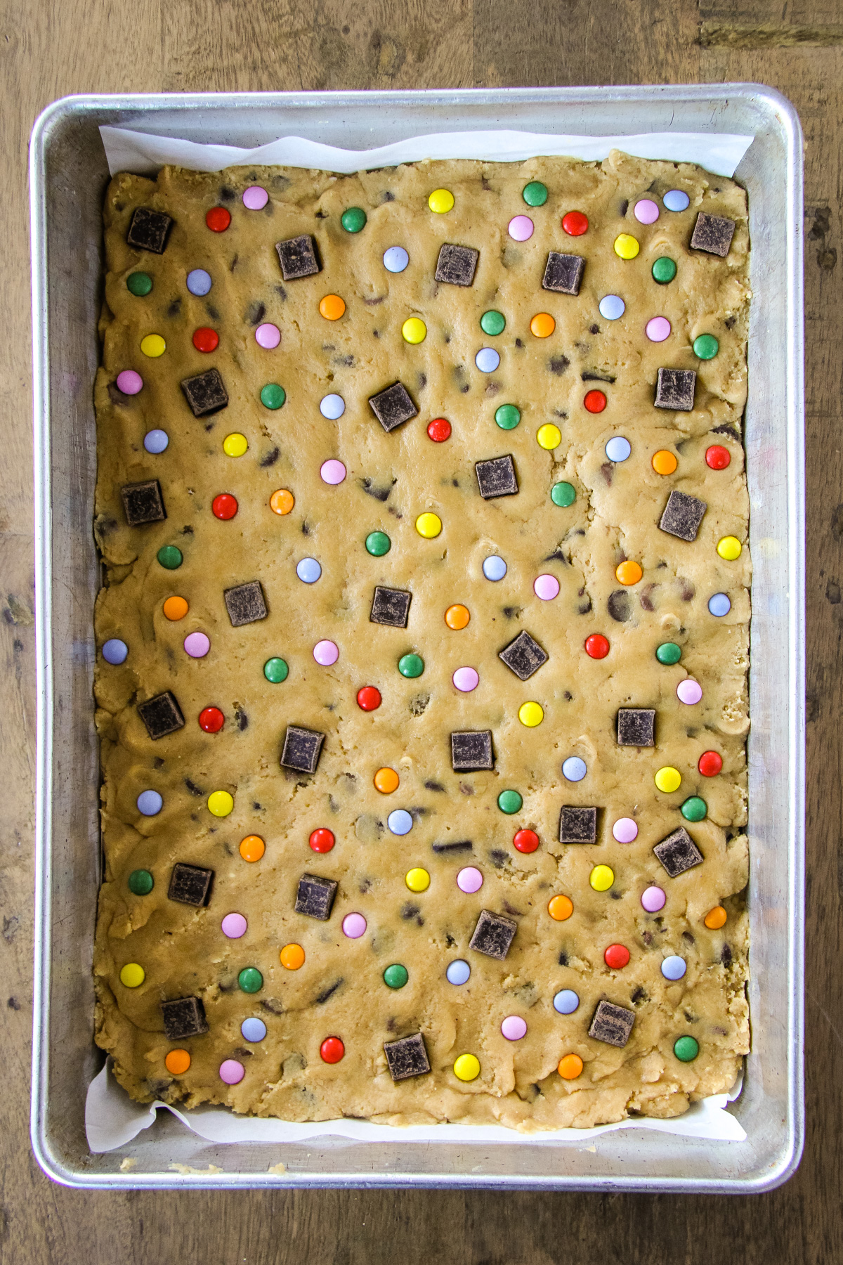a top down image of a baking pan filled with chocolate chip cookie dough dotted with coloured chocolate candy