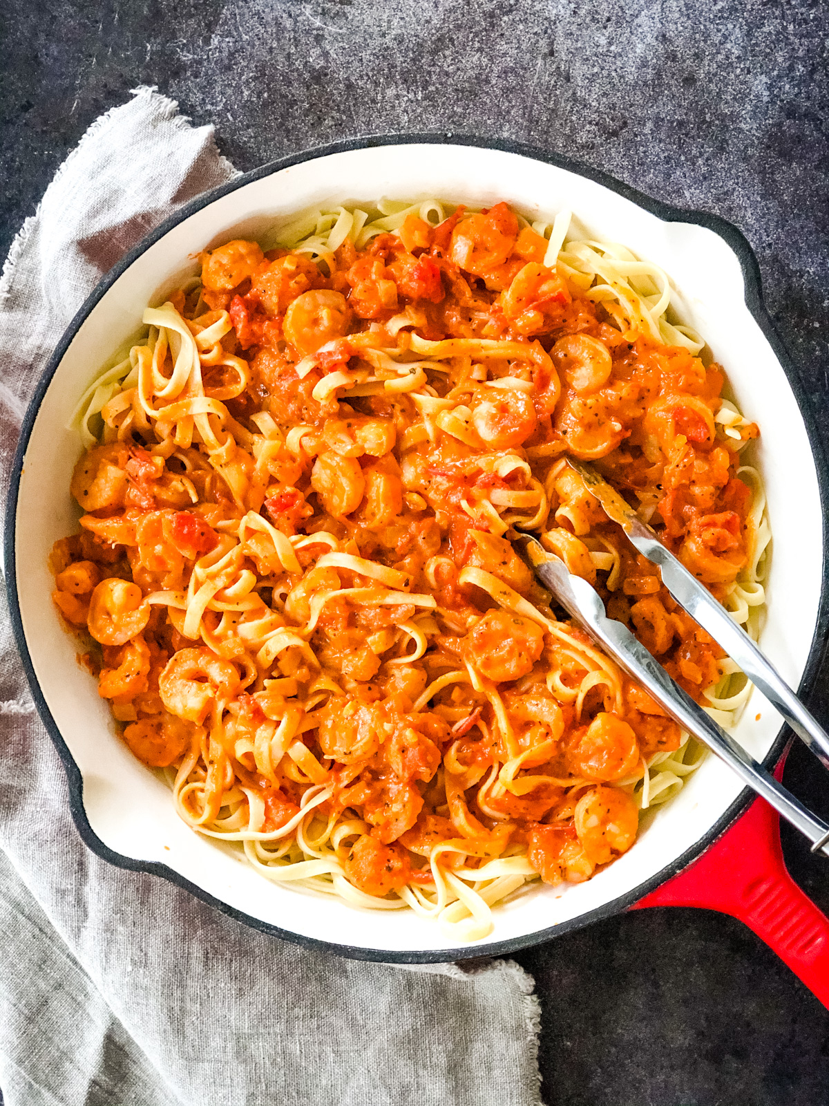 a top down image of a dish of tomato basil shrimp pasta with a set of tongs mixing