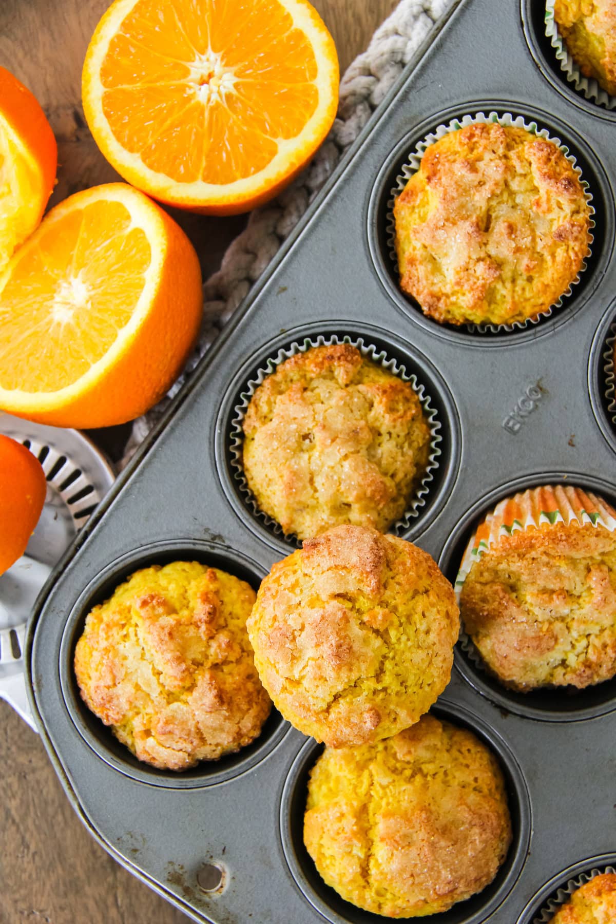 a top down image of a muffin tin filled with orange blender muffins. next to the tin are two orange halves