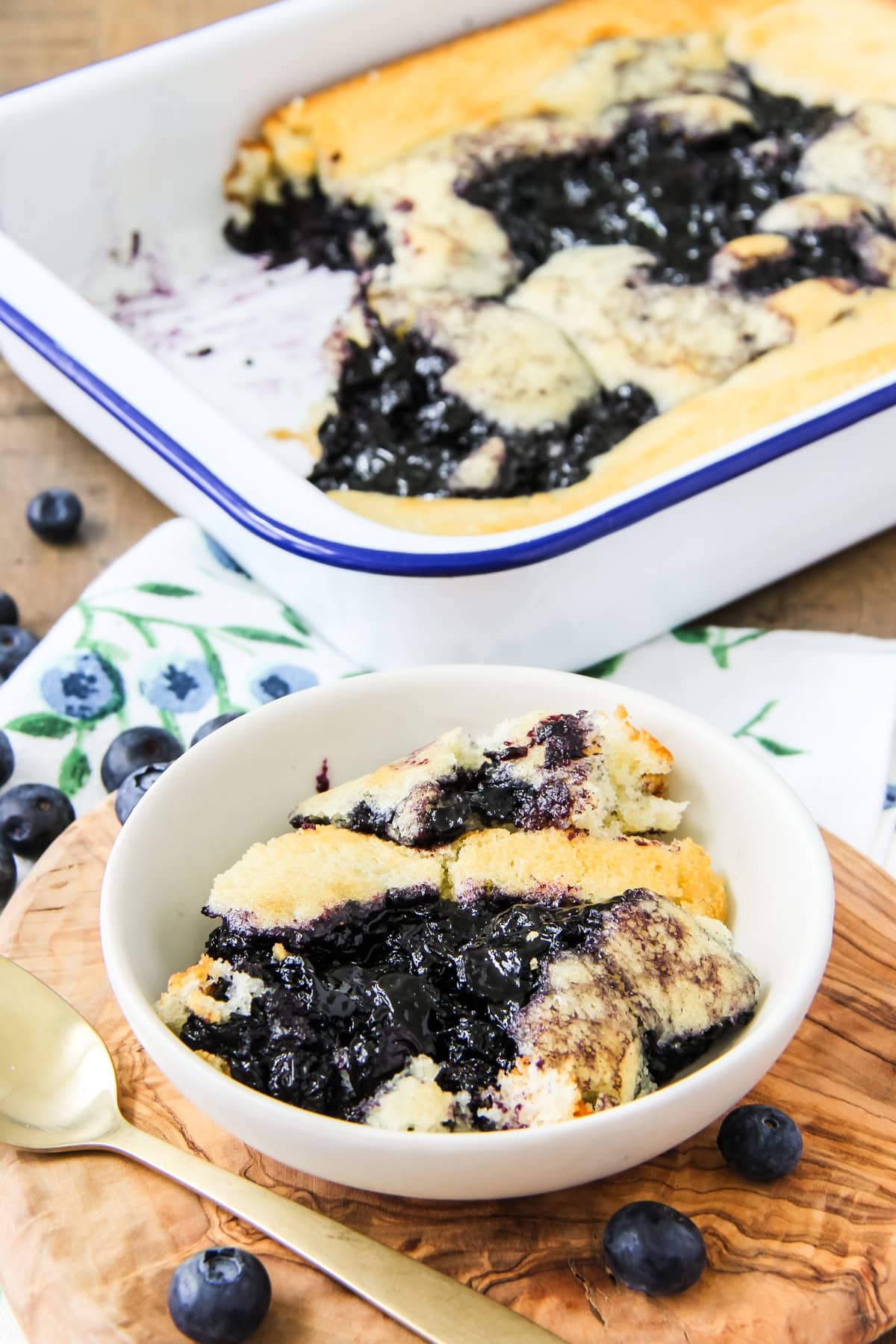 a scoop of blueberry pudding cake in a white bowl sitting next to a pan of pudding cake