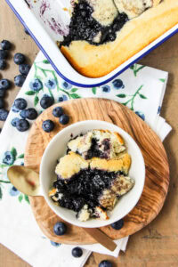 a scoop of blueberry pudding cake in a white bowl sitting next to a pan of pudding cake
