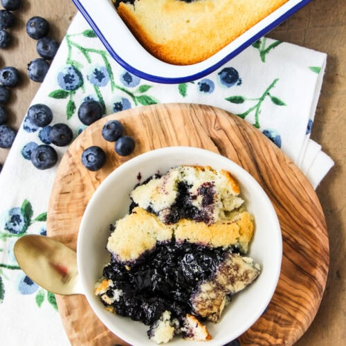 a scoop of blueberry pudding cake in a white bowl sitting next to a pan of pudding cake